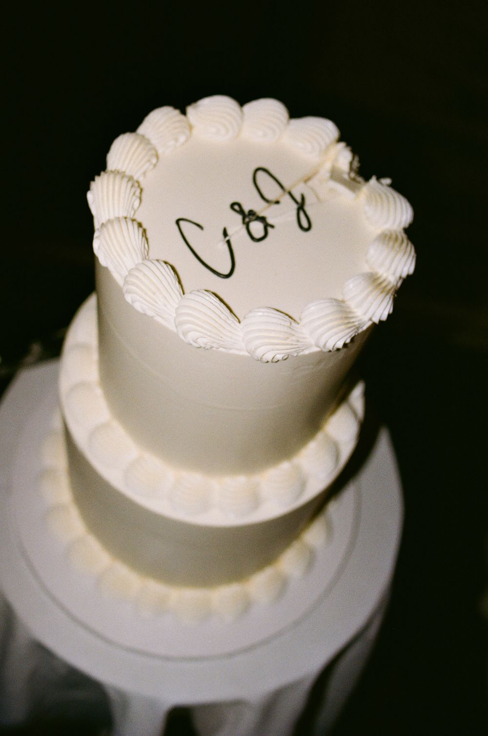 Two-tier white wedding cake with scalloped frosting and C&J monogram on top.
