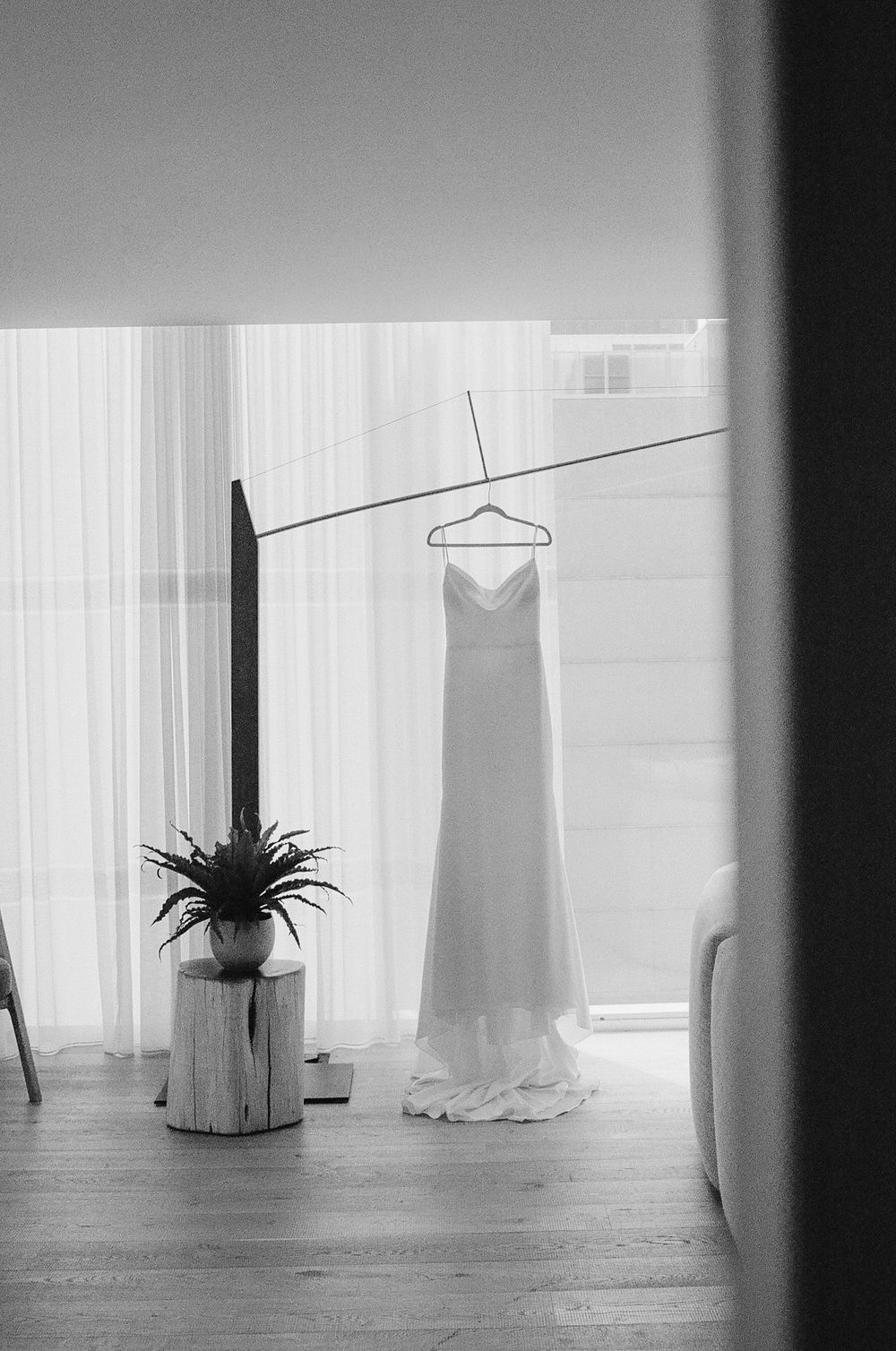Black and white photo of an elegant wedding dress hanging on a rod near sheer curtains with a plant on a wooden stump.