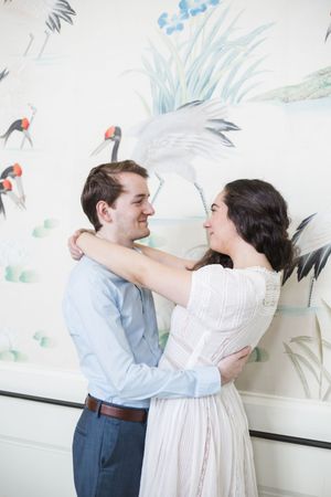 Couple in light colored outfits dancing and smiling against a wall with bird artwork.