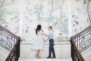 A romantic engagement photoshoot on an elegant staircase with floral wallpaper backdrop.