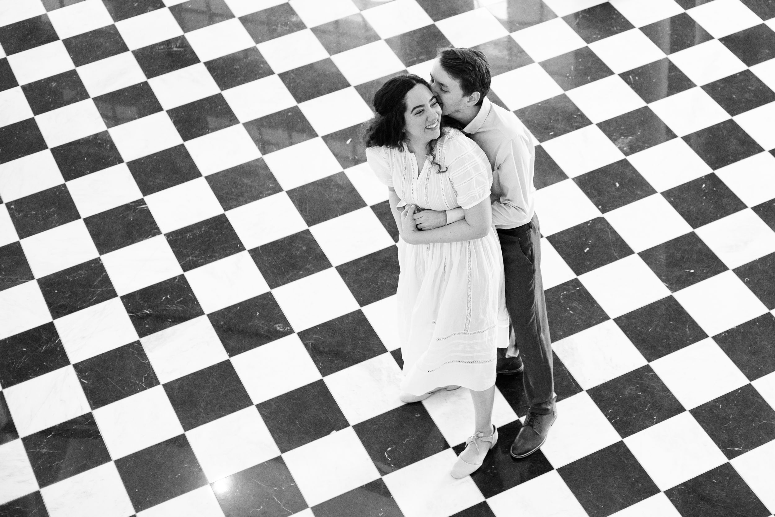 Two people in white clothing sit on a black and white checkered floor in a creative artistic pose.