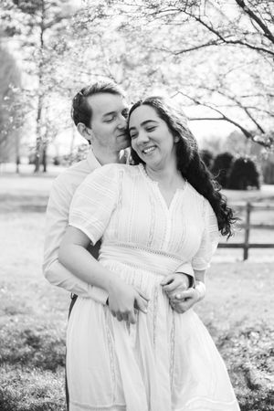 Romantic couple in white clothing share tender moments under blooming cherry blossom trees in a park setting.