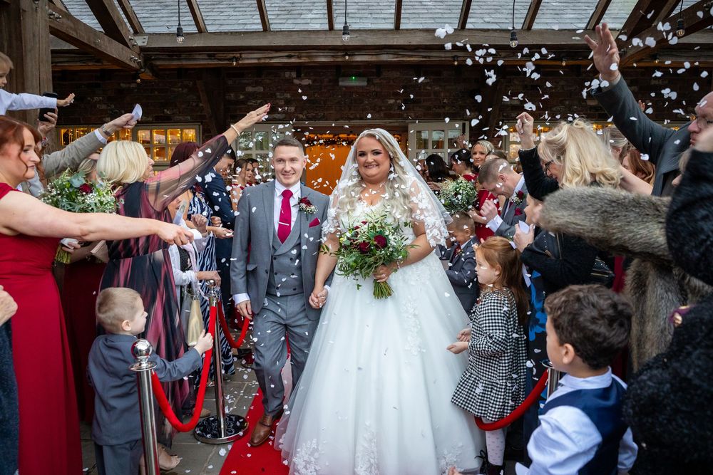 Bride and groom walk through confetti shower on red carpet surrounded by cheering guests at Charnock Farm venue.