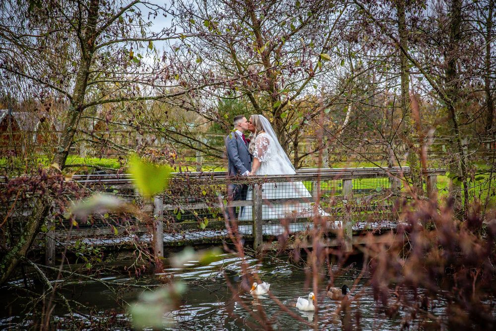 Bride and groom share a romantic kiss on a bridge over a pond surrounded by lush greenery.