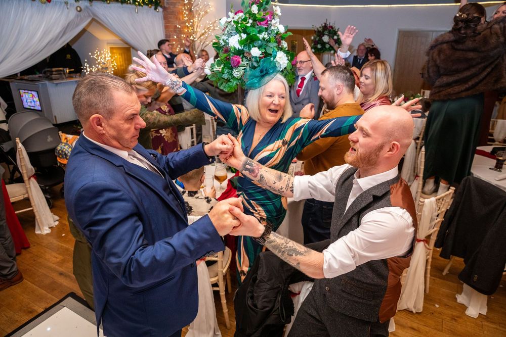 Joyful wedding guests dance and celebrate together near a decorated Christmas tree indoors.