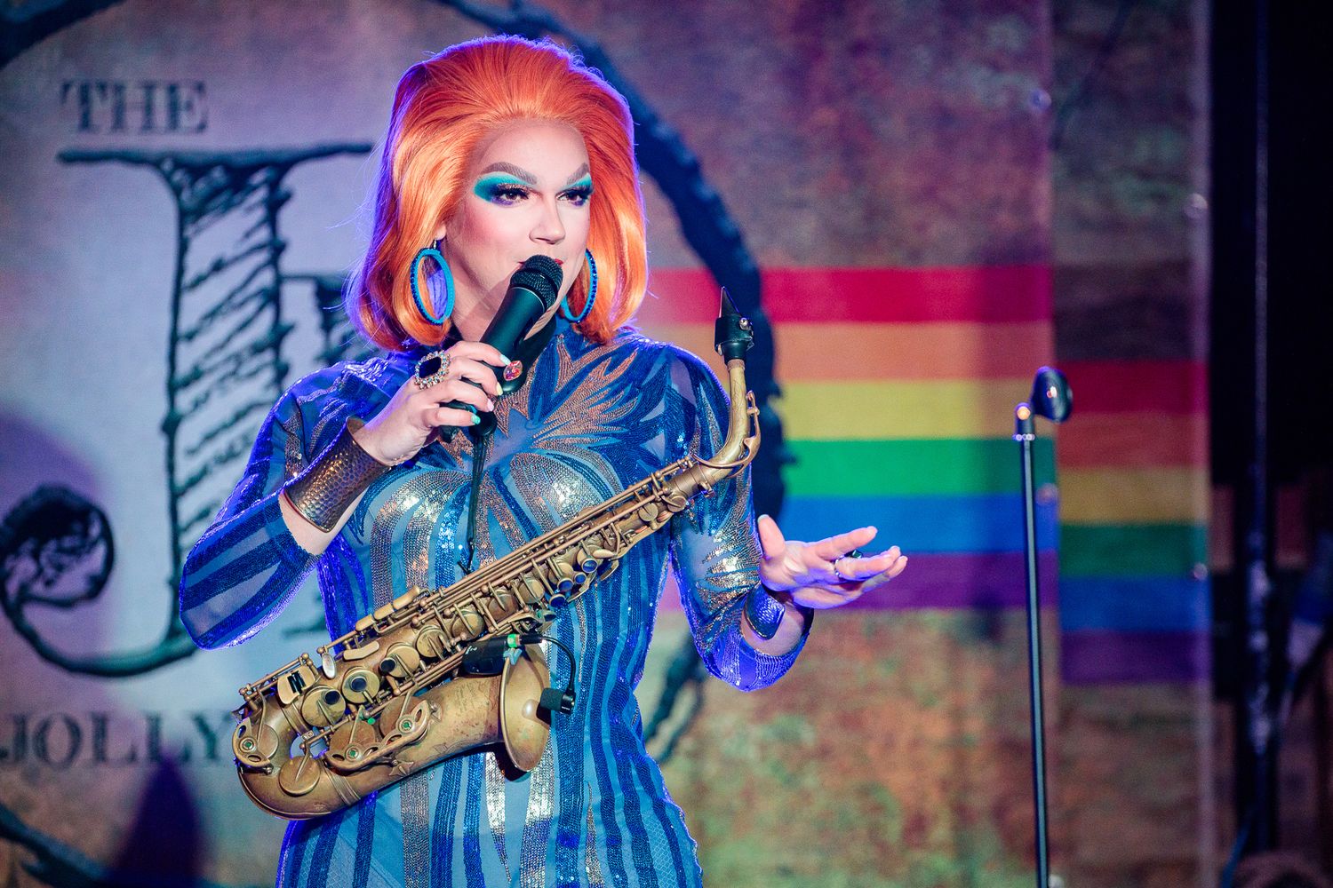A performer in a blue dress plays saxophone on stage with colorful lighting in the background.
