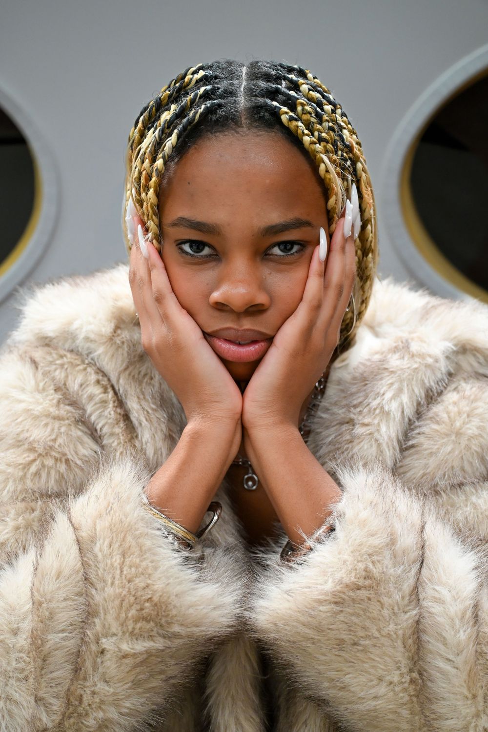 Fashion portrait of a model in a beige fur coat with blonde braids posing with hands on face against gray background.