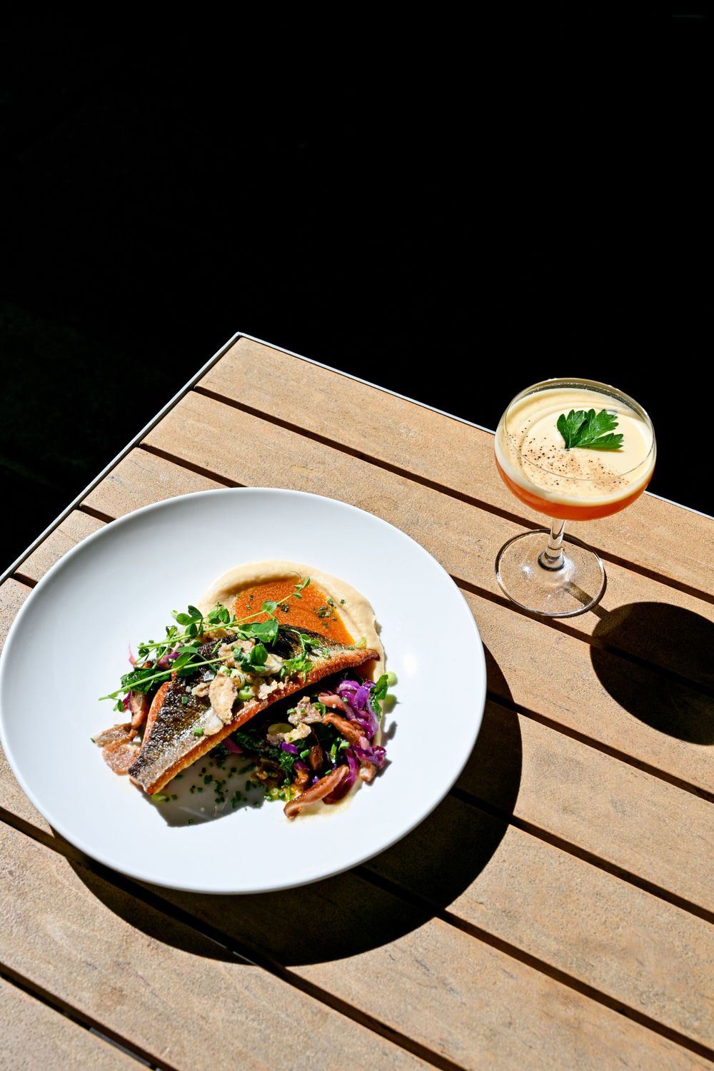Elegantly plated fish dish with cocktail on wooden outdoor table in sunlight.