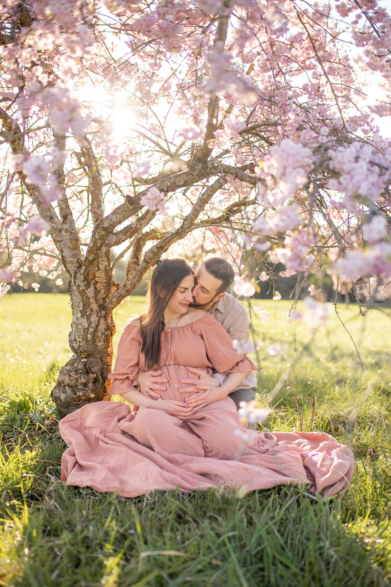 Femme enceinte et son compagnon enlacés sous un arbre en fleurs au printemps dans les Yvelines