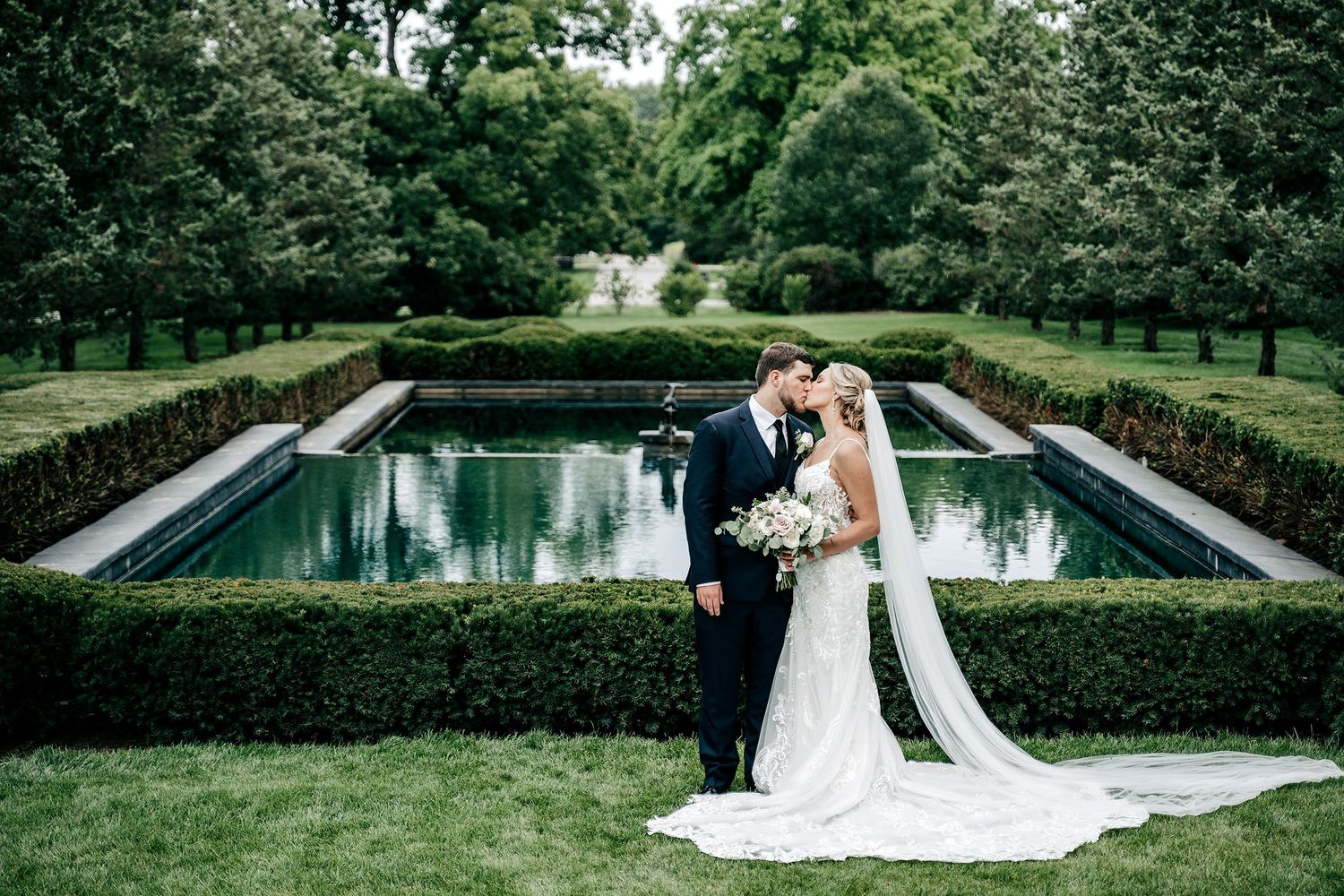 A romantic wedding moment captured beside a formal garden pool surrounded by manicured hedges and lush greenery.