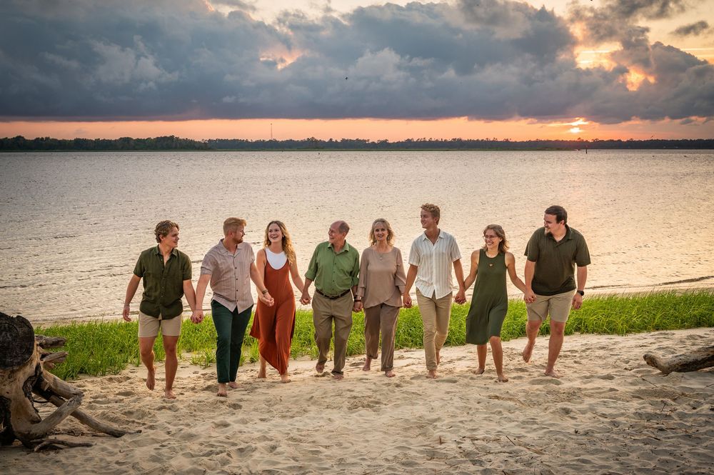 A group walks together along a sandy beach at sunset with dramatic clouds and a calm lake in the background.