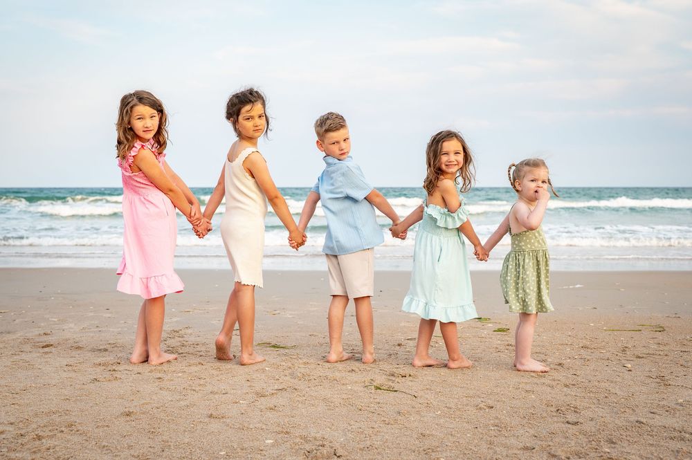 Children in pastel dresses hold hands while playing on sandy beach at sunset.