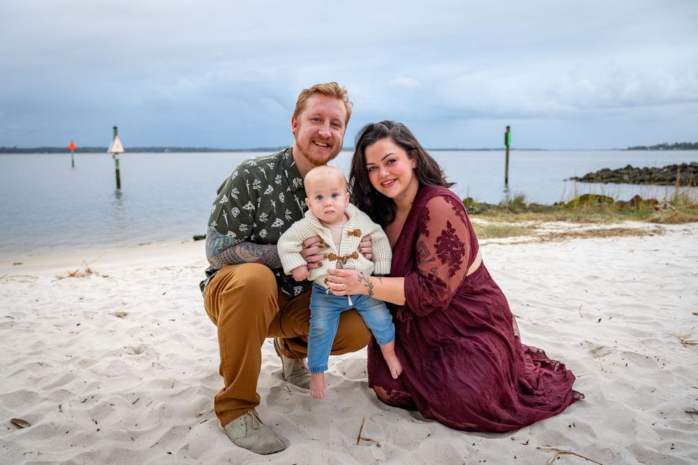 Family of three poses together on a sandy beach with water markers and cloudy sky visible behind them.