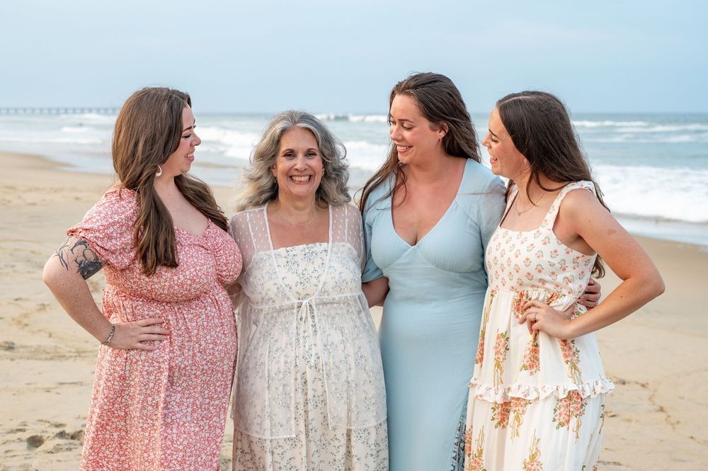 A group in coordinating floral and pastel dresses stands together on a sunny beach by the ocean.