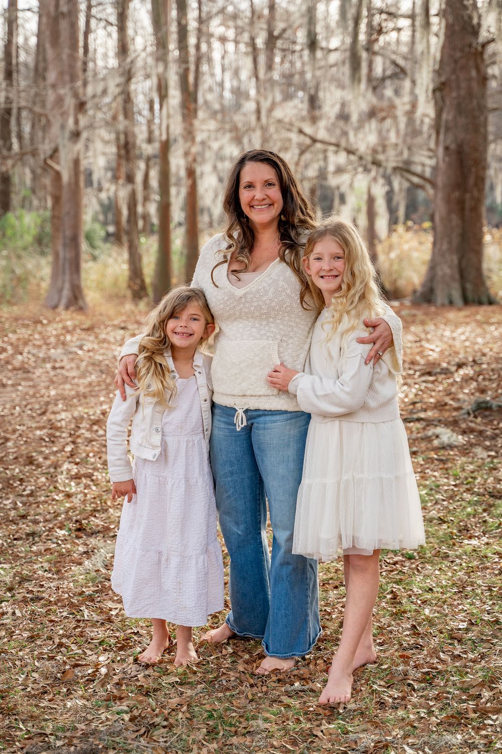 A family group poses together on fallen autumn leaves in a wooded setting wearing coordinated white and neutral outfits.