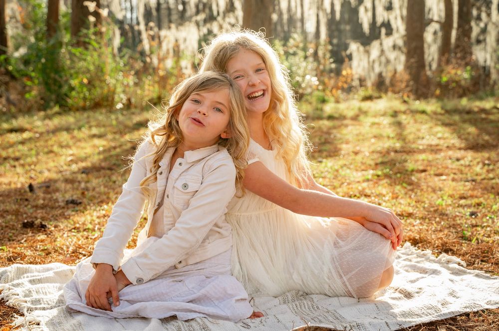 Two people in white dresses sit together on a blanket in a sun-dappled forest setting during golden hour.