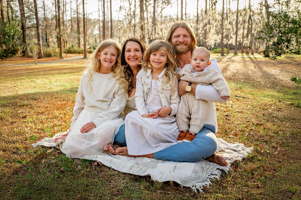 Family members wearing white and denim sit together on a blanket in a sunlit autumn forest setting.