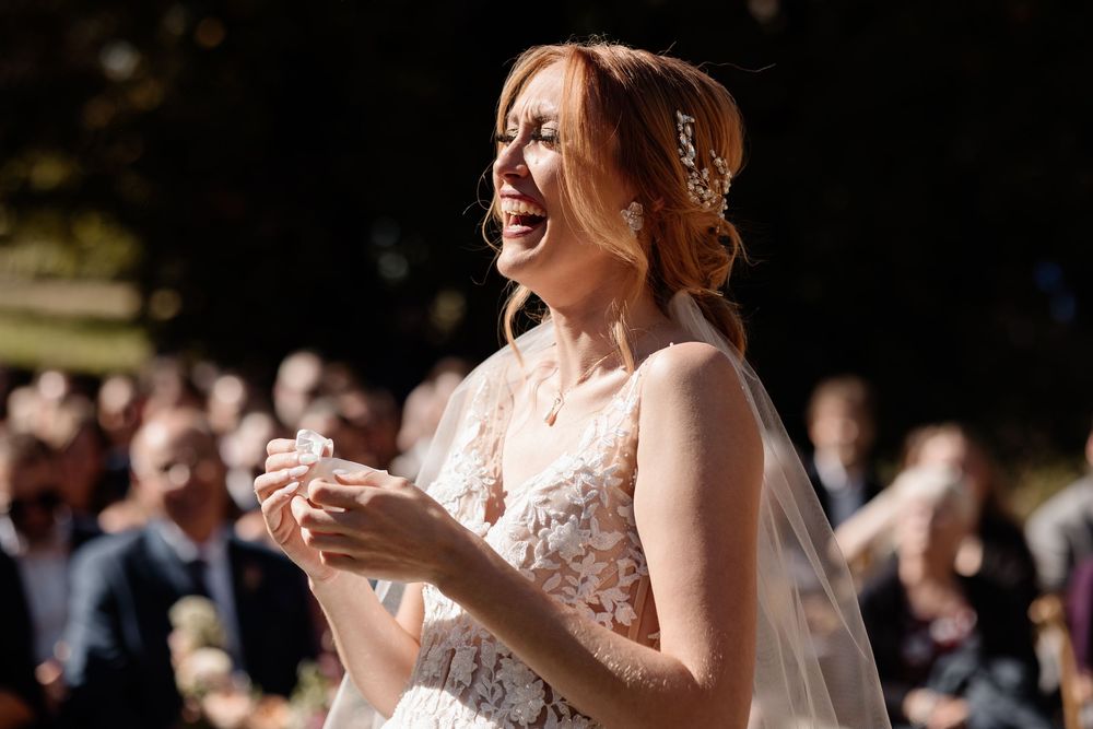 Emotional bride in lace wedding dress and veil, laughing with joy during outdoor ceremony surrounded by guests. Candid Wedding Photography.