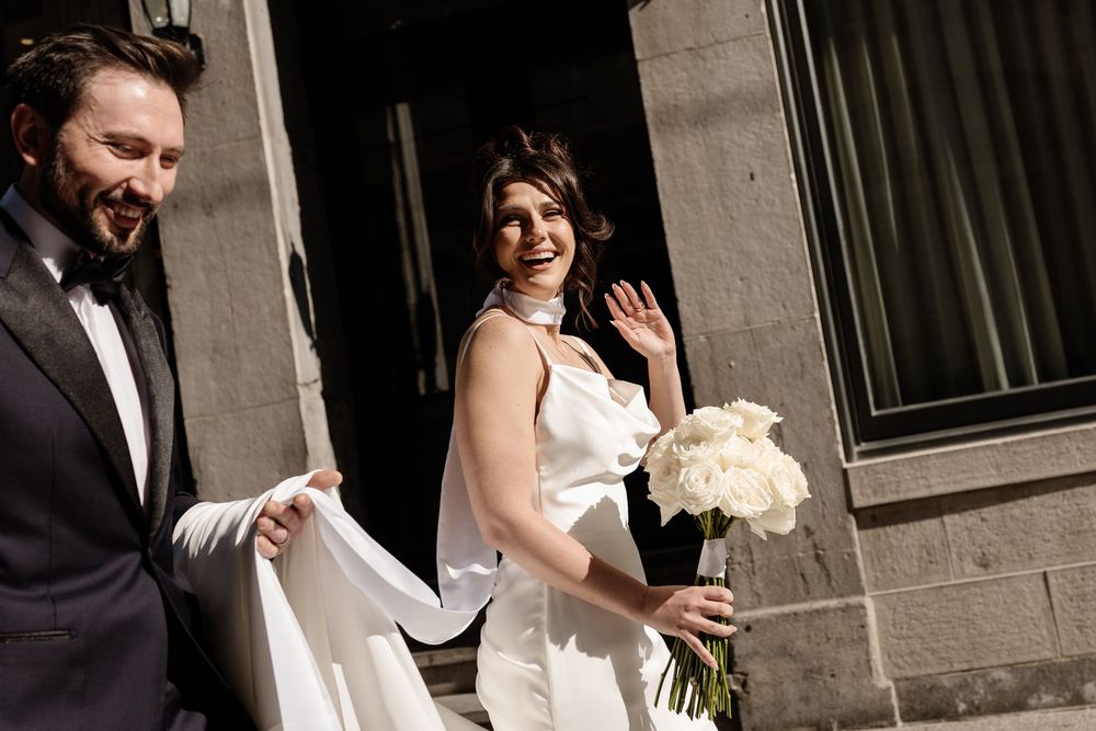 Smiling bride in white satin gown holds white peony bouquet beside groom in dark suit in Old Montreal. Candid Montreal Wedding Photography.