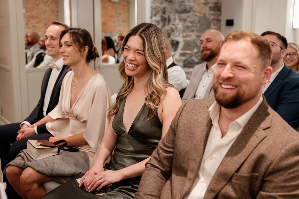 Guests laughing and smiling during a wedding ceremony seated in a bright white venue.