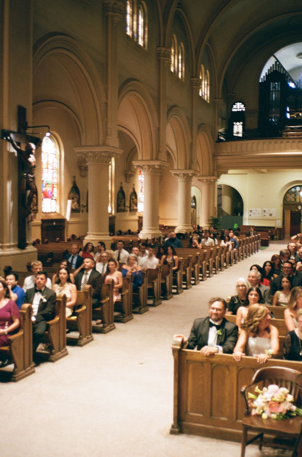 Film photos of guests in wooden pews, waiting for the bride, inside a grand Montreal church during a wedding ceremony with arched ceilings.
