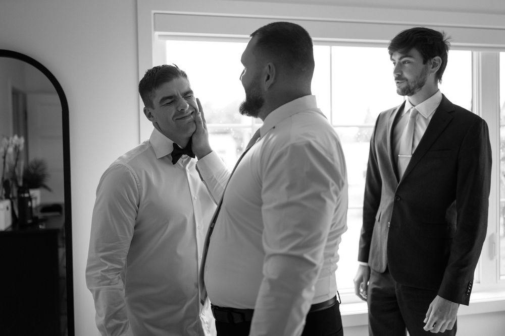 Black and white photo of three men in formal attire conversing indoors before a wedding ceremony.