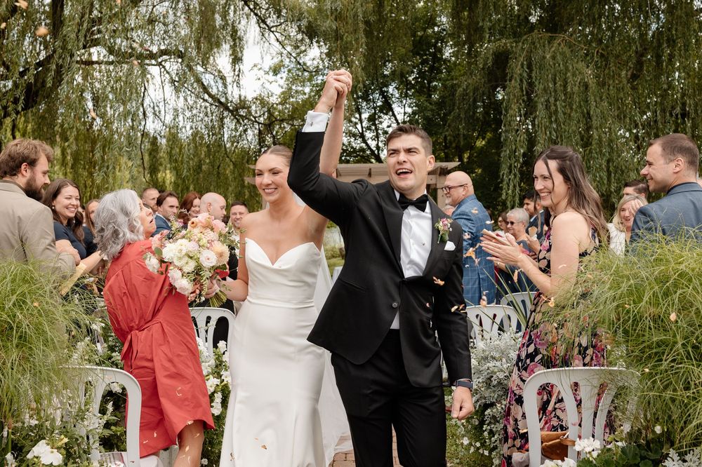 Joyful newlyweds walking down outdoor aisle surrounded by applauding guests under weeping willow trees. Candid Montreal Wedding Photographer