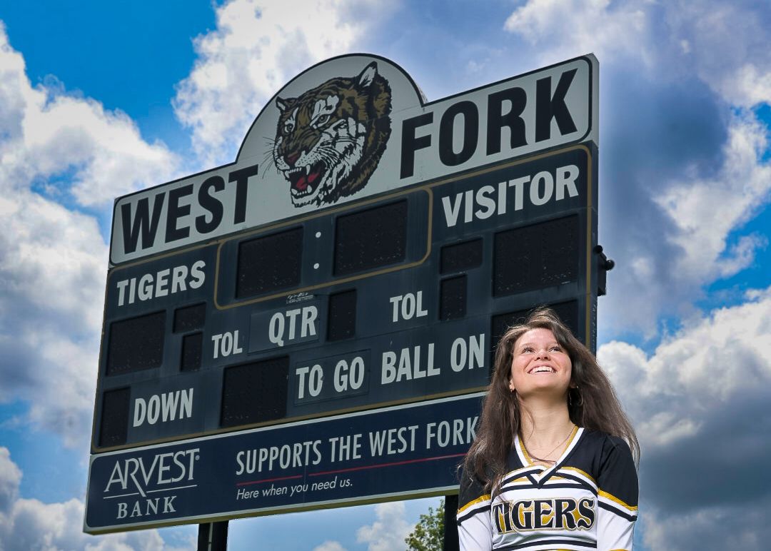 Cheerleader poses in front of West Fork Tigers scoreboard sponsored by Arvest Bank under a cloudy blue sky.