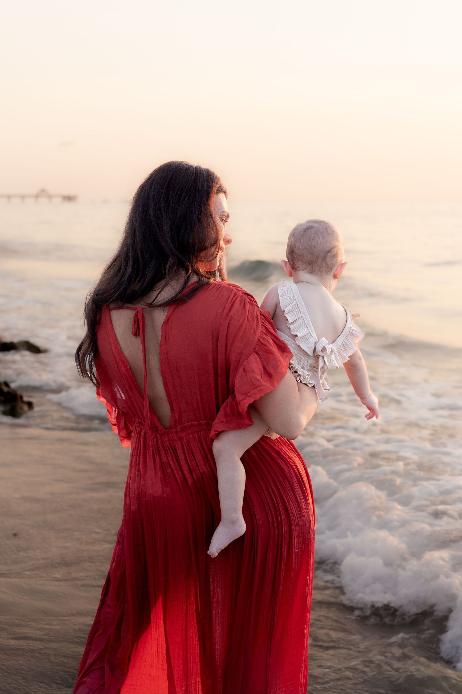 Woman in red dress holding baby on beach at sunset.