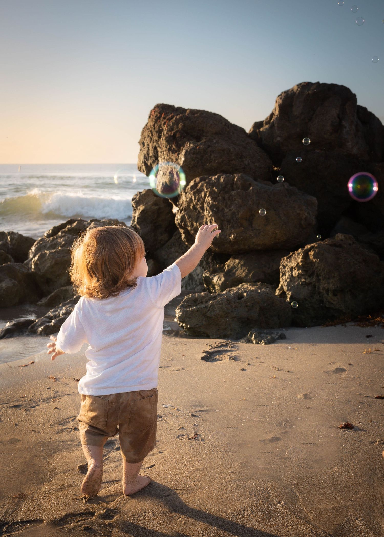 A little boy in brown shorts is blowing bubbles and chasing them at the beach at golden hour.