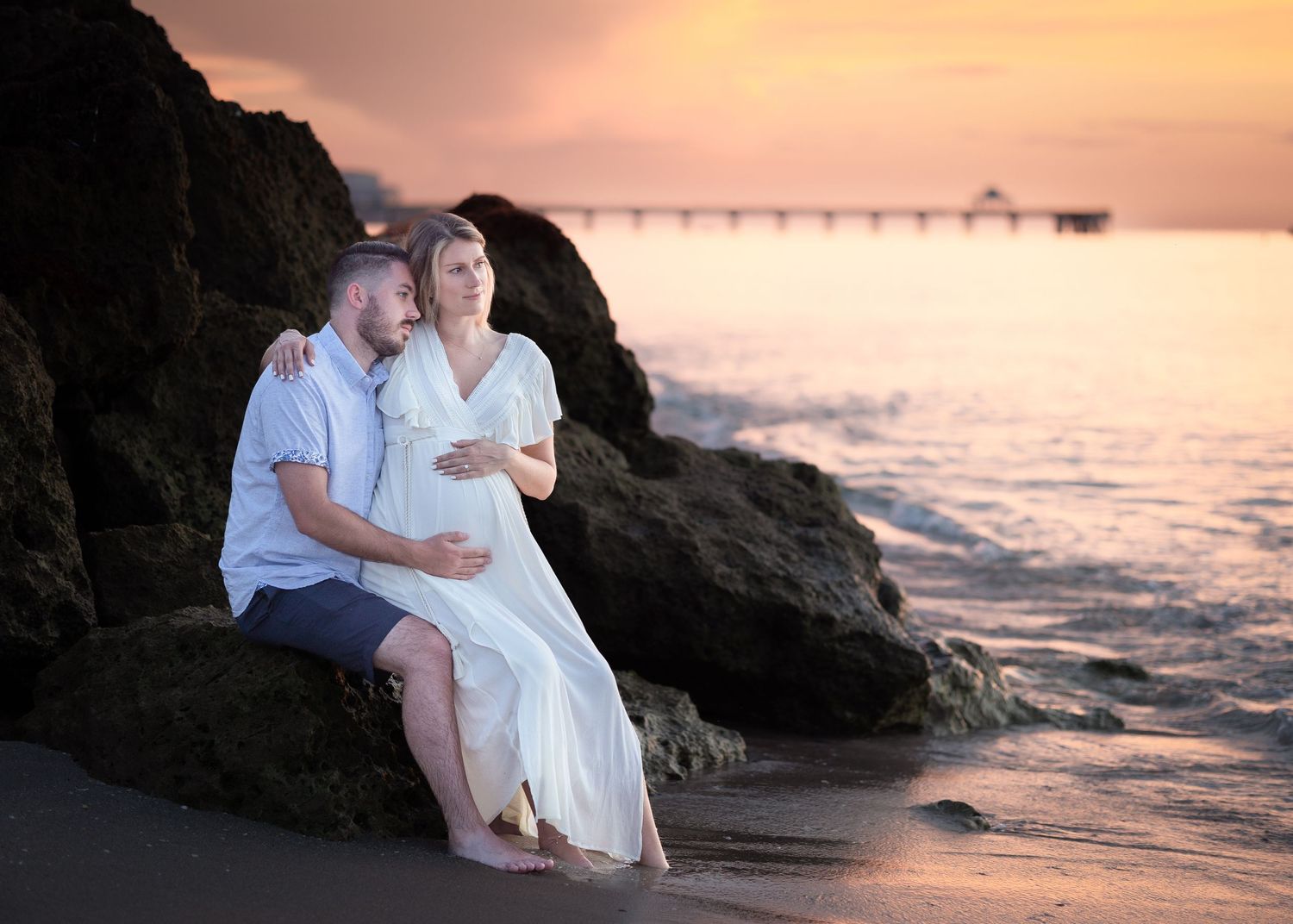 Couple sitting on beach rocks at sunset, pregnant woman in white dress, man embracing her with the Deerfield Beach pier in background.