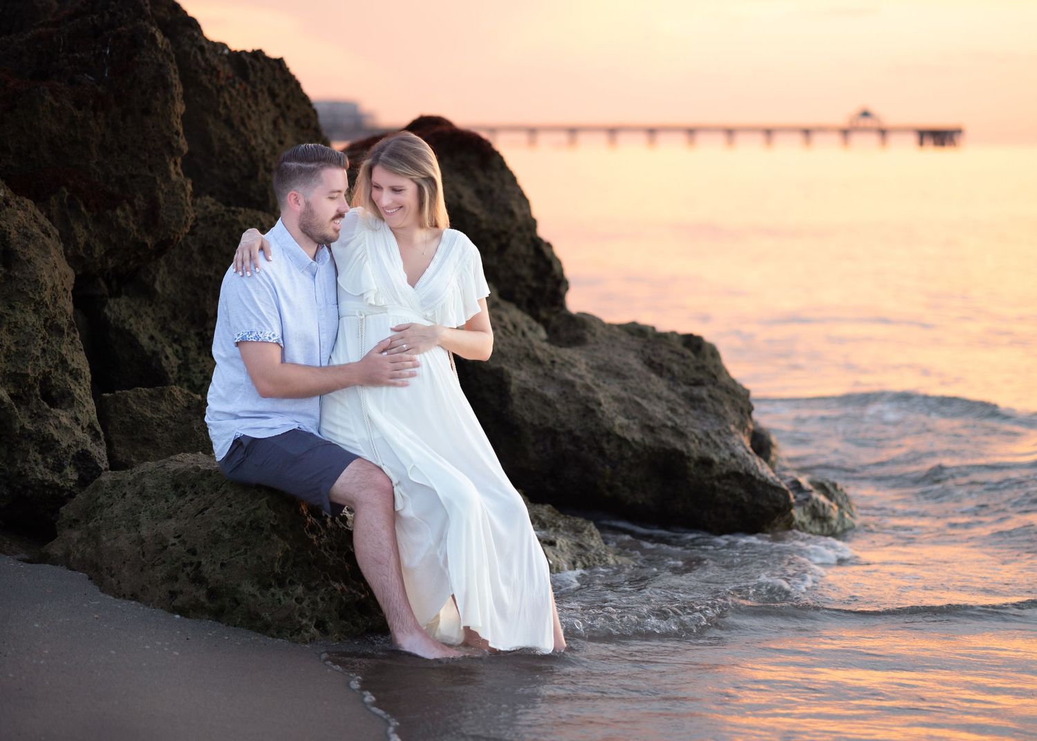 A pregnant woman and her partner are sitting on the rocks at the beach with the pier behind them at sunrise. He is holding her baby bump.