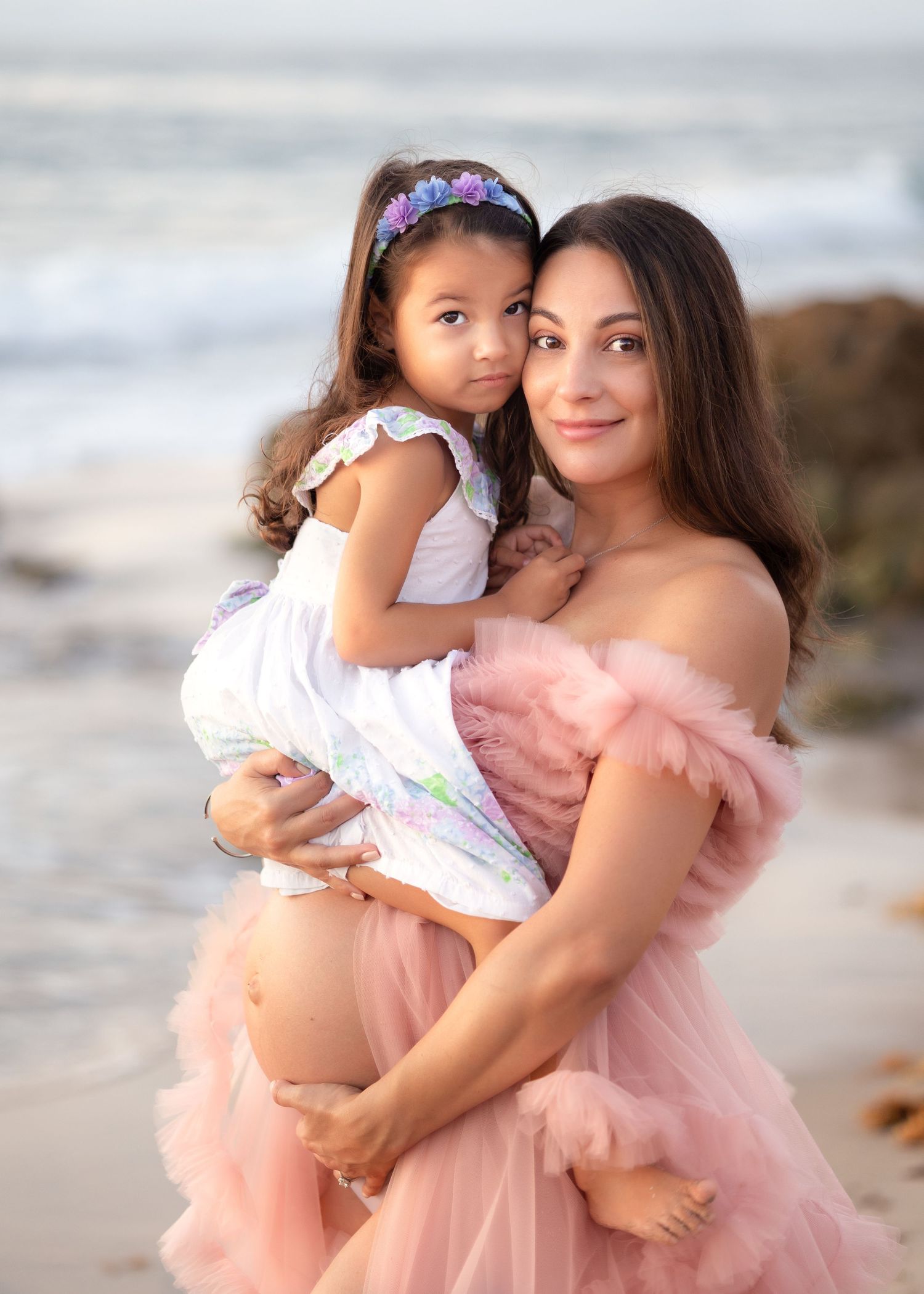 Pregnant woman in pink tulle gown holds young girl on beach, both smiling softly near ocean waves.