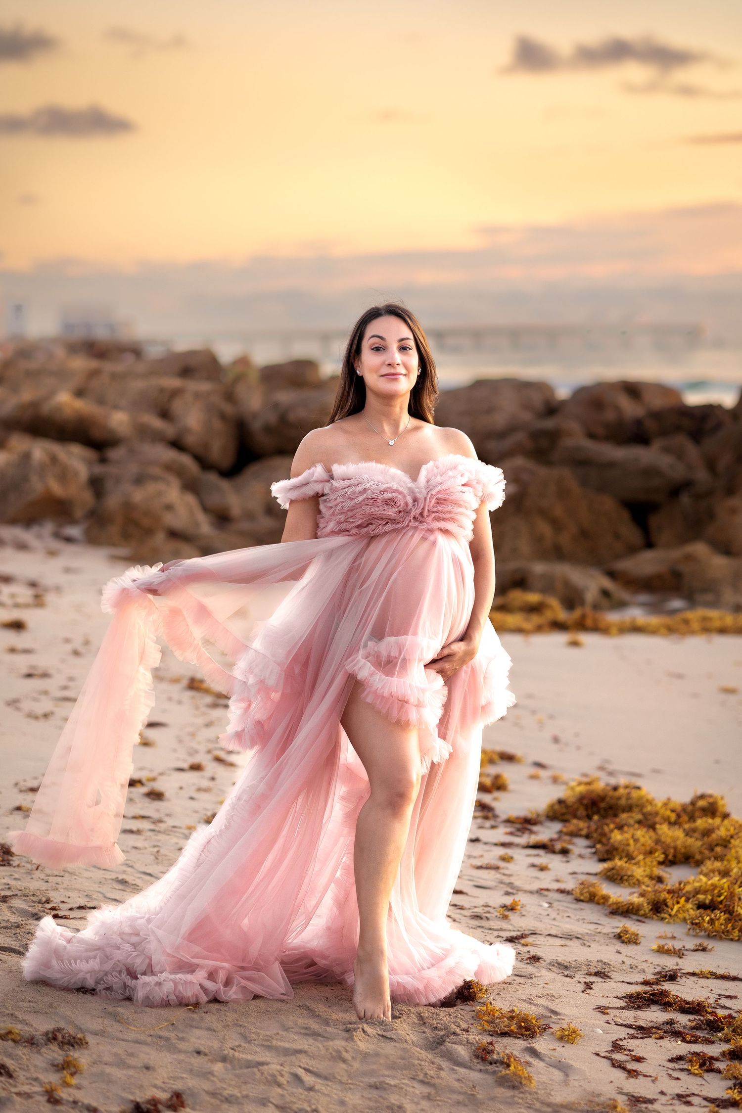Pregnant woman in flowing pink tulle gown poses on sandy beach at sunset with rocky shore backdrop.