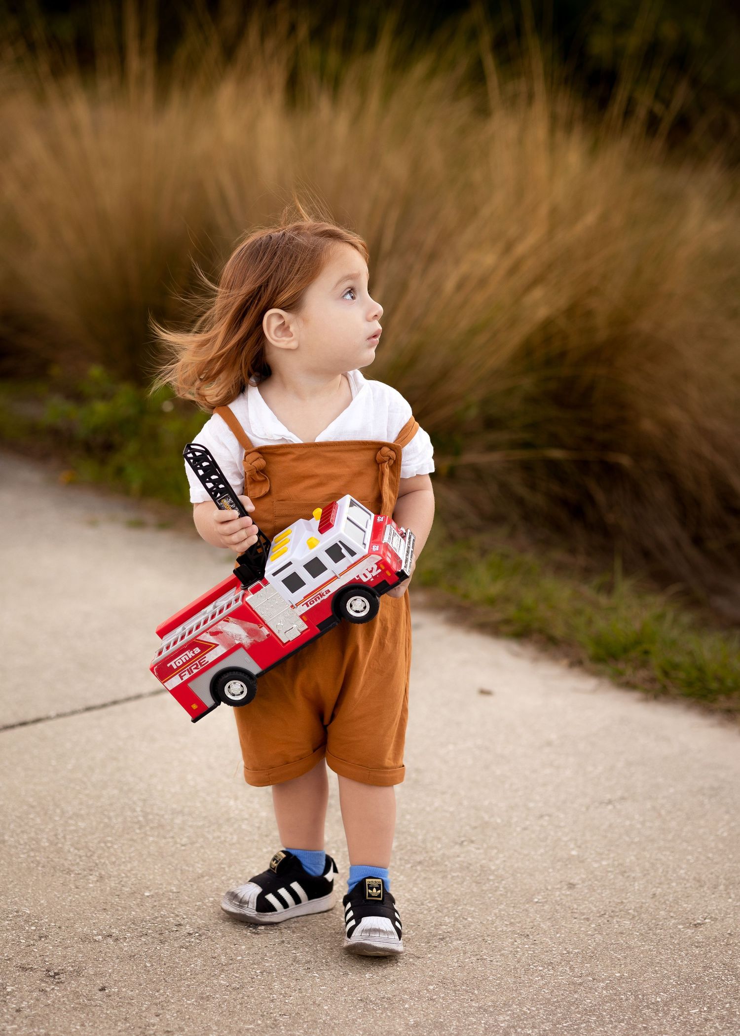 Toddler in brown overalls holding a toy fire truck on a park path, with windswept hair and black sneakers.