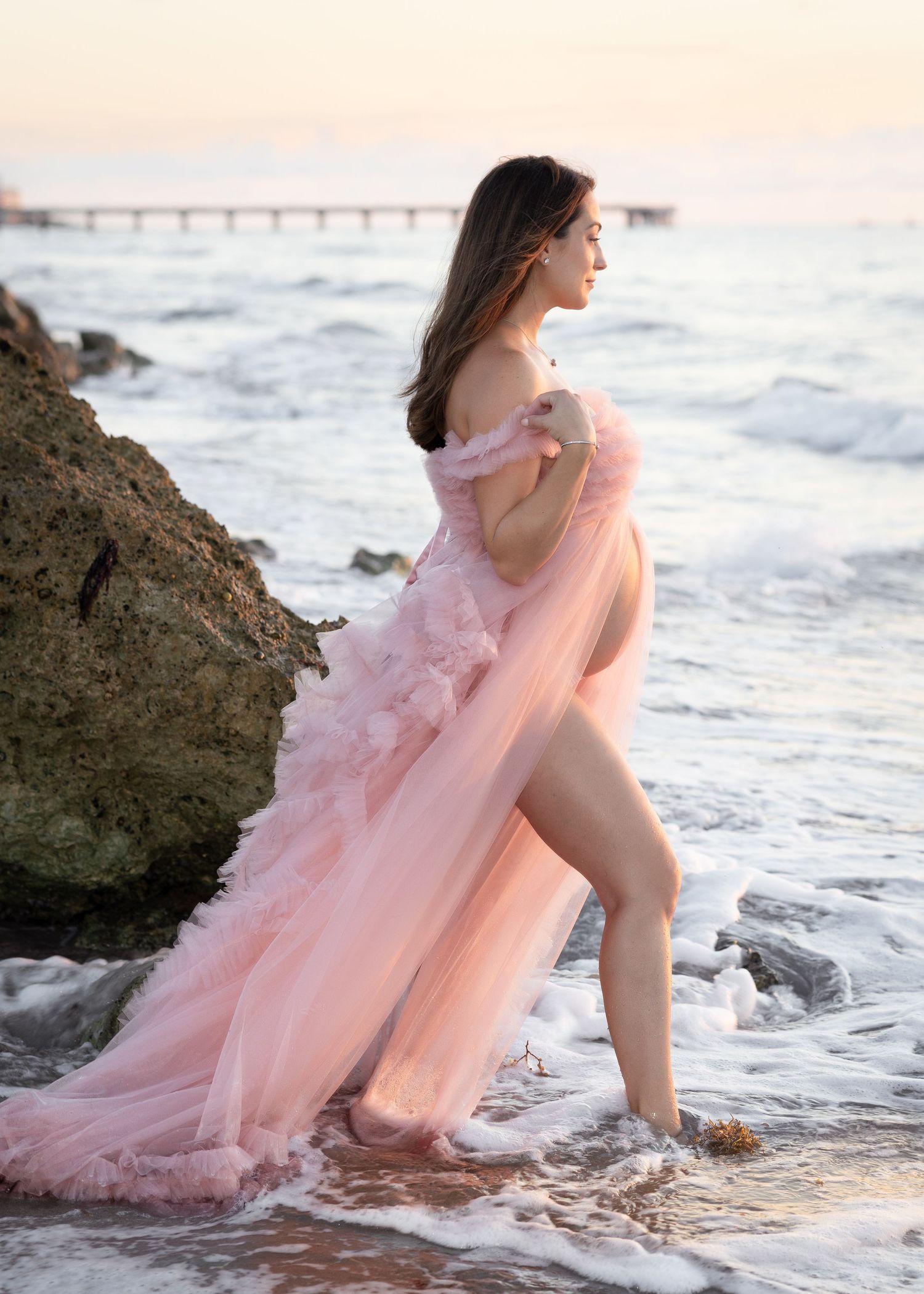 A pregnant woman in a flowing pink tulle gown off-shoulder gown stands on rocky shoreline at Deerfield Beach at sunset with ocean waves.