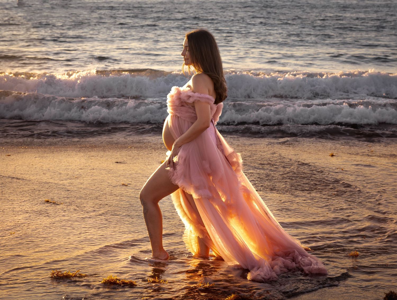 Pregnant woman in pink tulle gown poses on beach at golden sunset, waves crashing behind her.