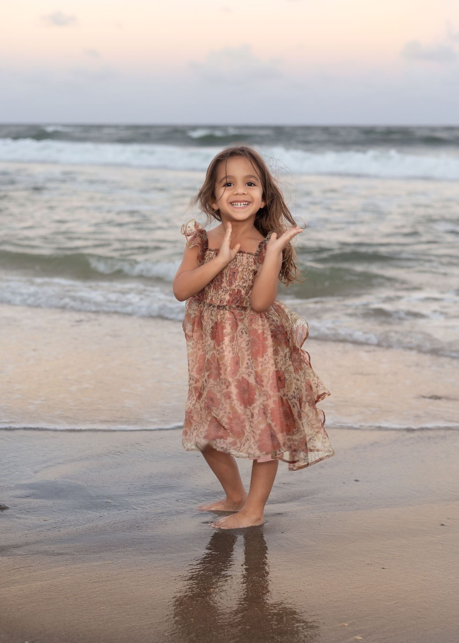 A young girl in a floral dress smiles and claps on a sandy beach with ocean waves behind her.
