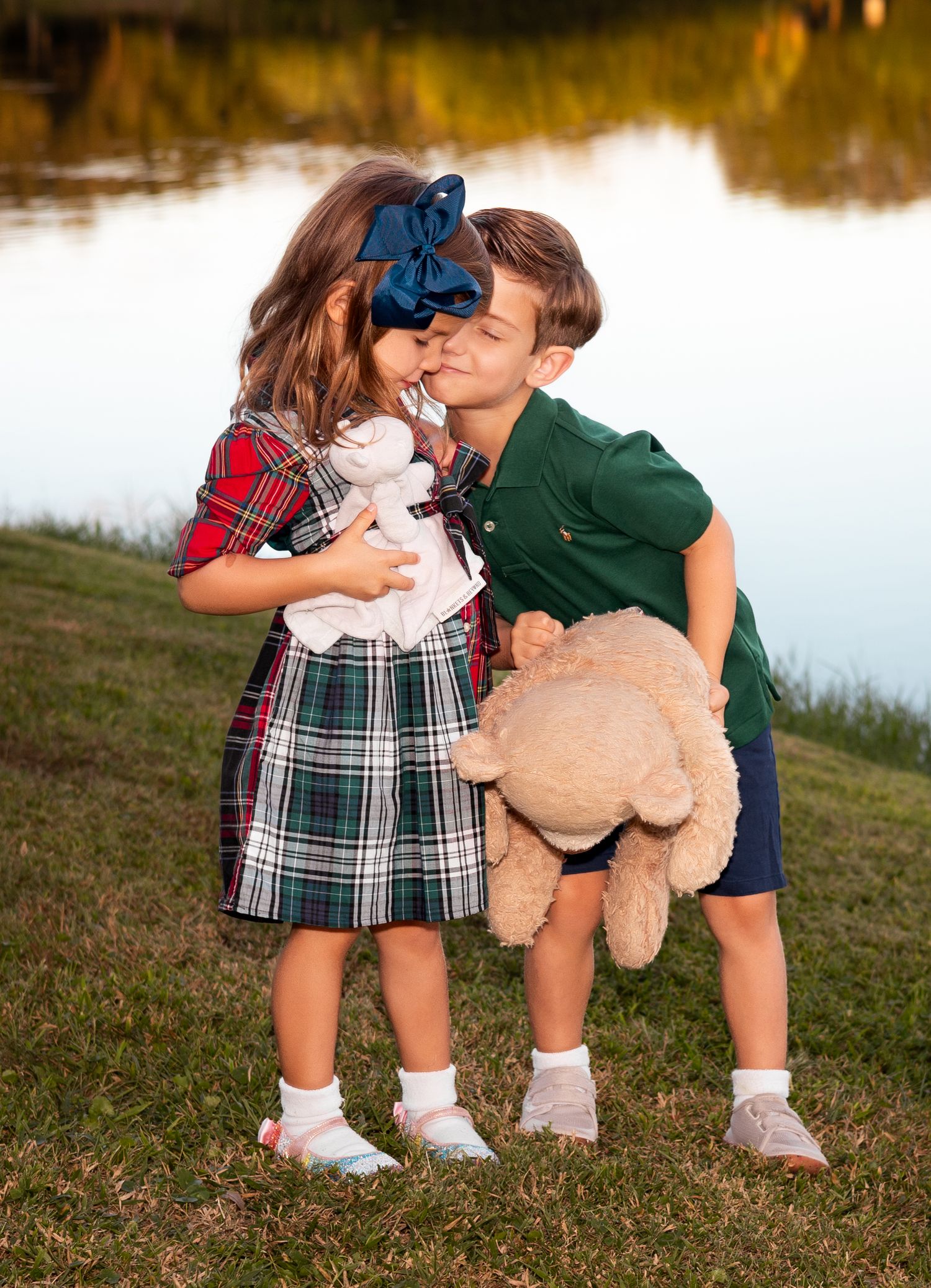 Two young children in plaid and green outfits share a sweet kiss while holding stuffed animals near a pond.