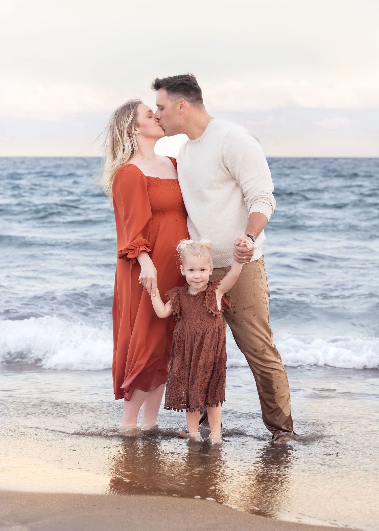A couple kisses on a beach while holding hands with their young daughter in matching earth-tone outfits near ocean waves.