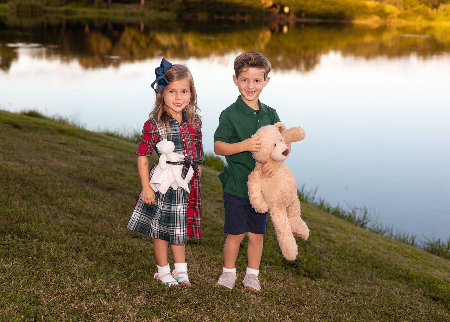 Two young children pose by a pond at sunset, holding stuffed animals, dressed in coordinating outfits.