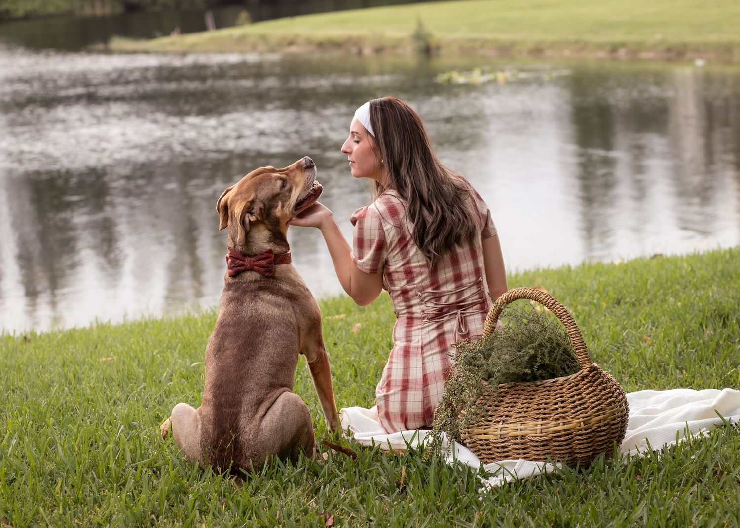Woman in plaid dress feeding her dog by a serene lake with a wicker basket at golden hour. Windjammer Park, FL.