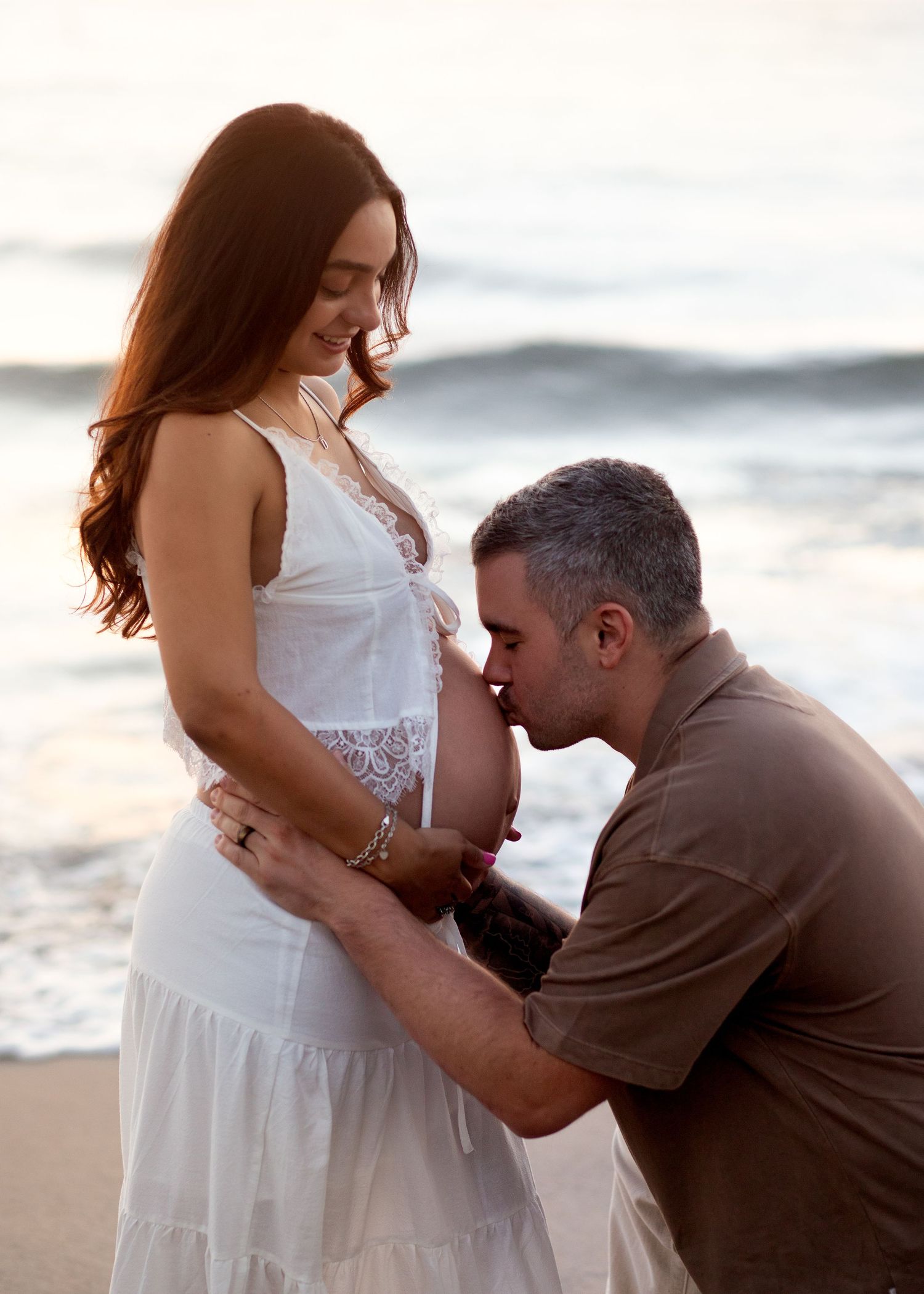 Man kisses pregnant woman's belly on beach at sunset, couple sharing tender maternity moment by ocean waves.