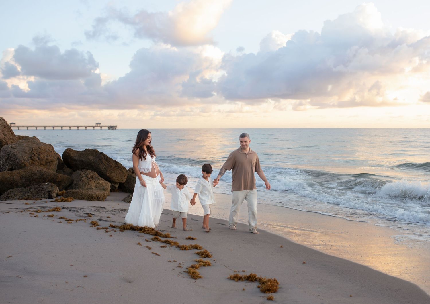 Family of four walking hand-in-hand on a sandy beach at sunset with ocean waves and a pier in the background.