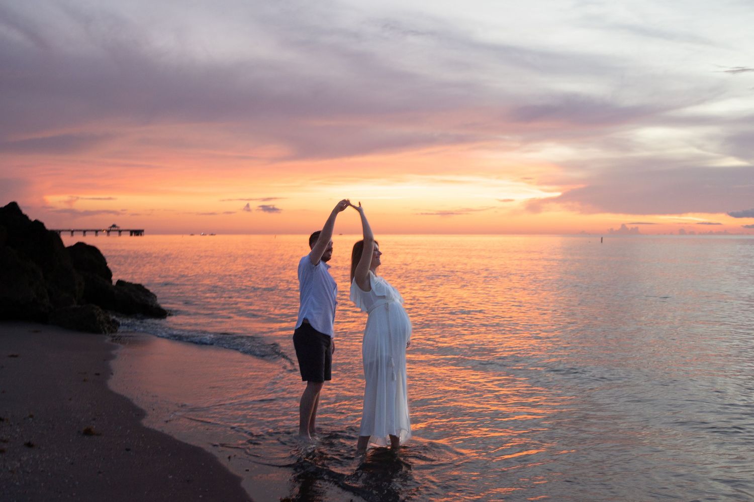 Couple dancing on a tropical beach at sunset, with vibrant pink and purple skies reflecting on calm ocean waters.