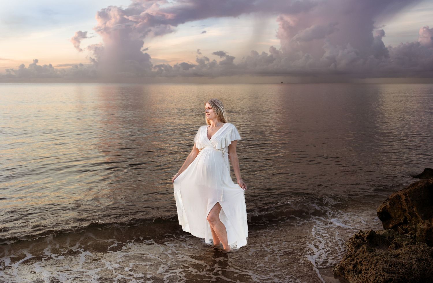 Woman pregnant woman in white dress standing on rocky shore at sunset with dramatic cloudy sky backdrop.