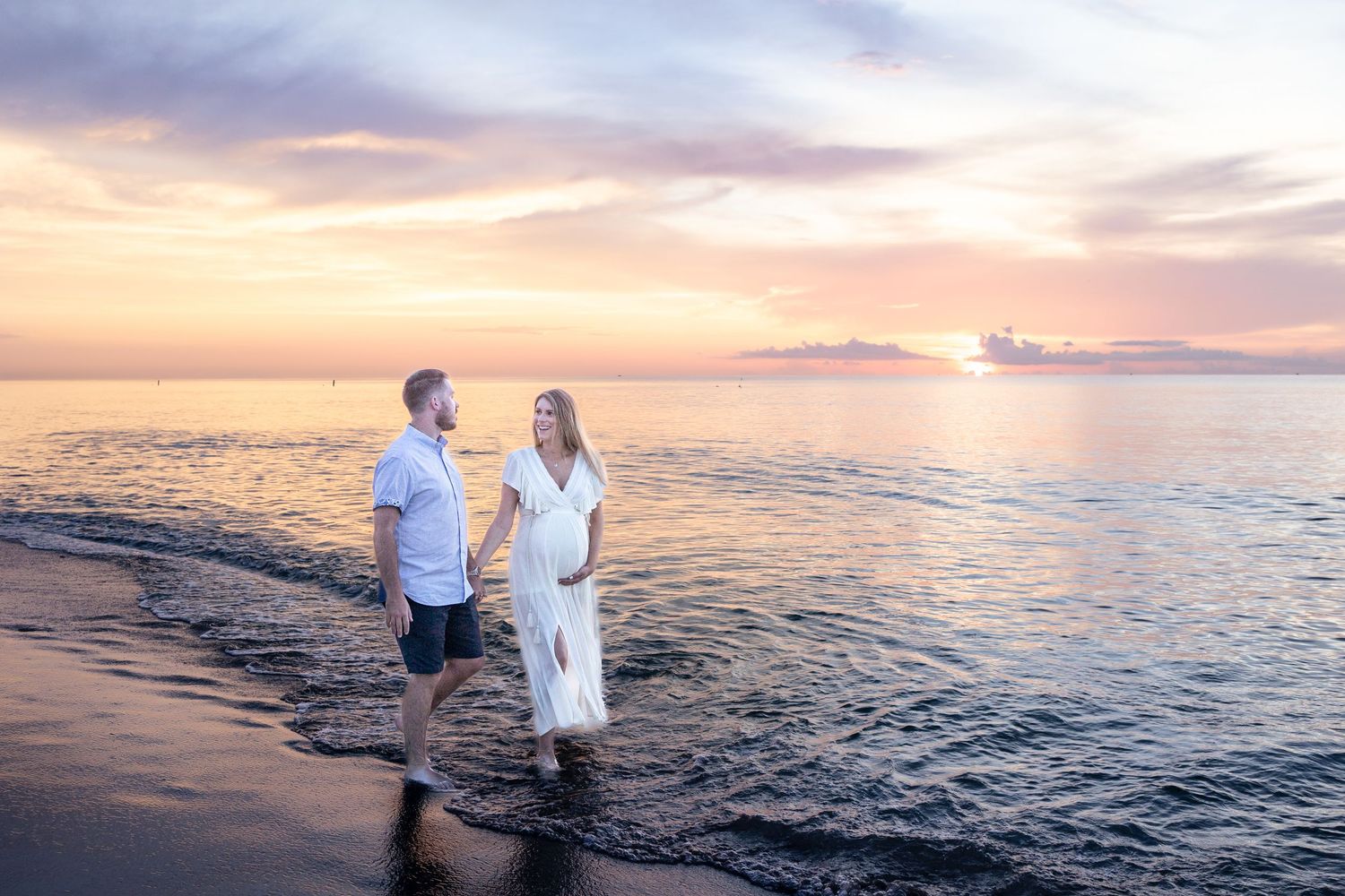A couple holds hands on a beach at sunset, the pregnant woman wearing a flowing white dress by the ocean.