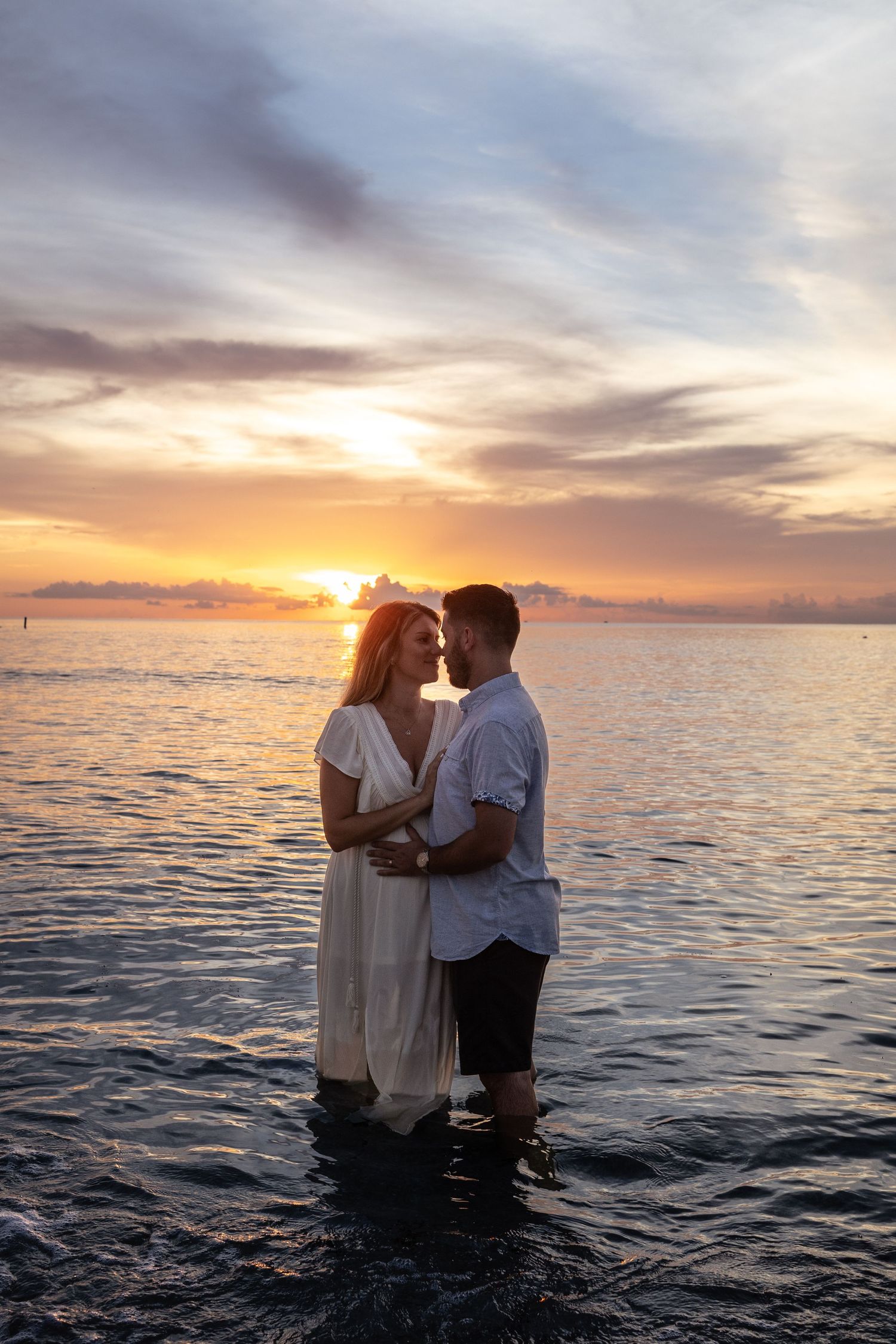 Couple standing in ocean water embracing at golden sunset, woman in white dress, romantic seaside moment.