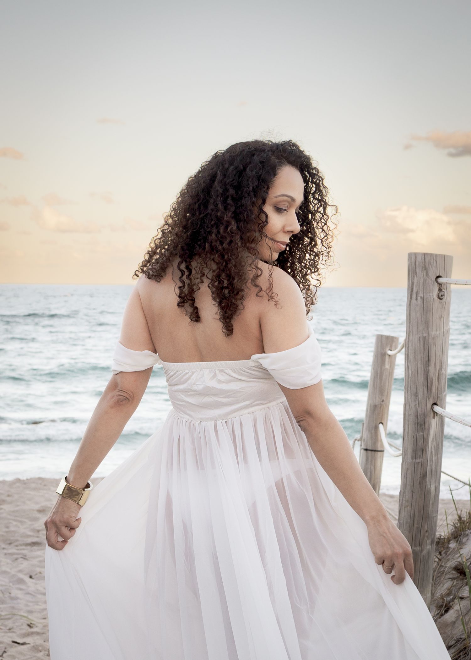 Woman in white off-shoulder dress posing on a beach at sunset with curly hair and gold bracelet.
