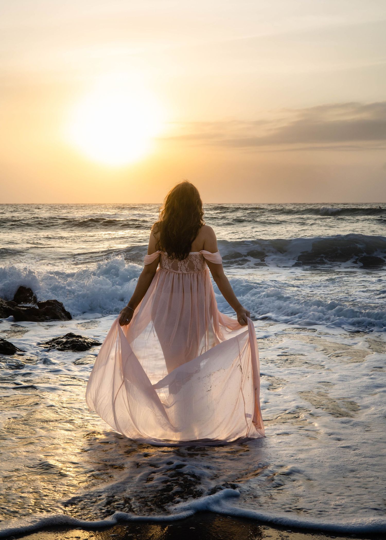 Woman in flowing pink gown standing at ocean's edge during golden sunset, waves crashing around her feet.